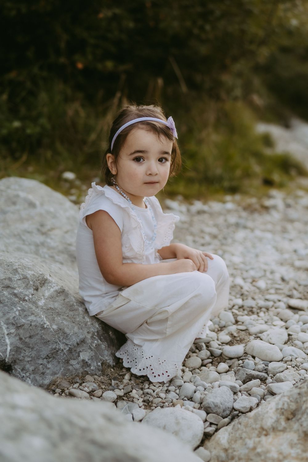 Young child in white outfit sitting on rocky terrain with a bow headband.