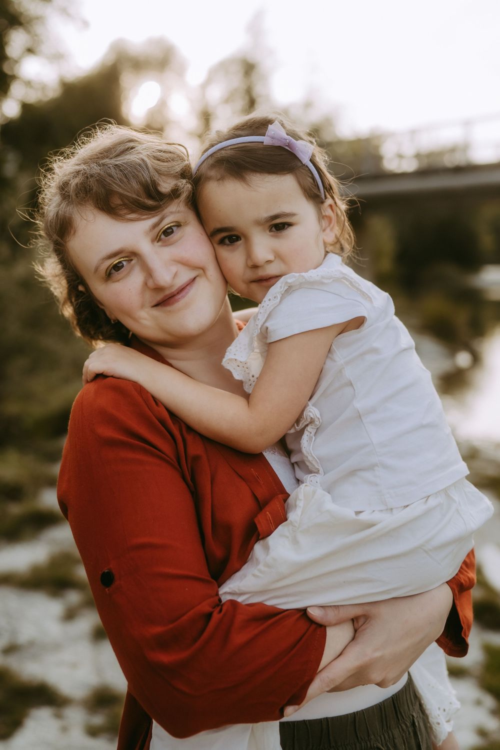 Parent in red sweater hugging small child in white dress outdoors.