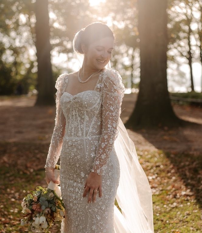 A bride in a lace long-sleeve wedding dress holds a pastel bouquet while standing in a sunlit forest setting.