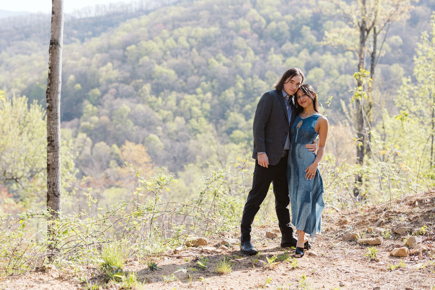 Couple embracing tenderly on a rocky hillside surrounded by spring foliage and rolling mountain views.