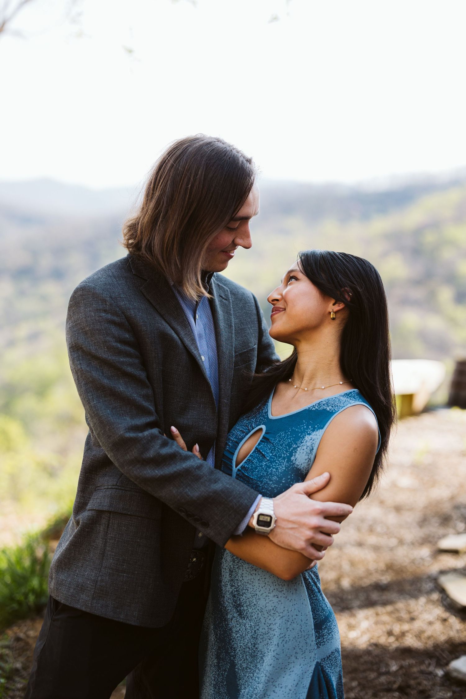 Couple gazing lovingly into each other eyes while embracing outdoors with a lush mountain valley behind them.