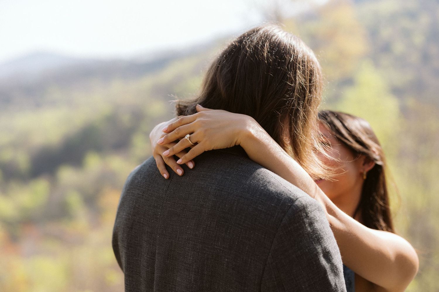 Close-up of woman embracing man from behind showing an engagement ring with mountain scenery in background.
