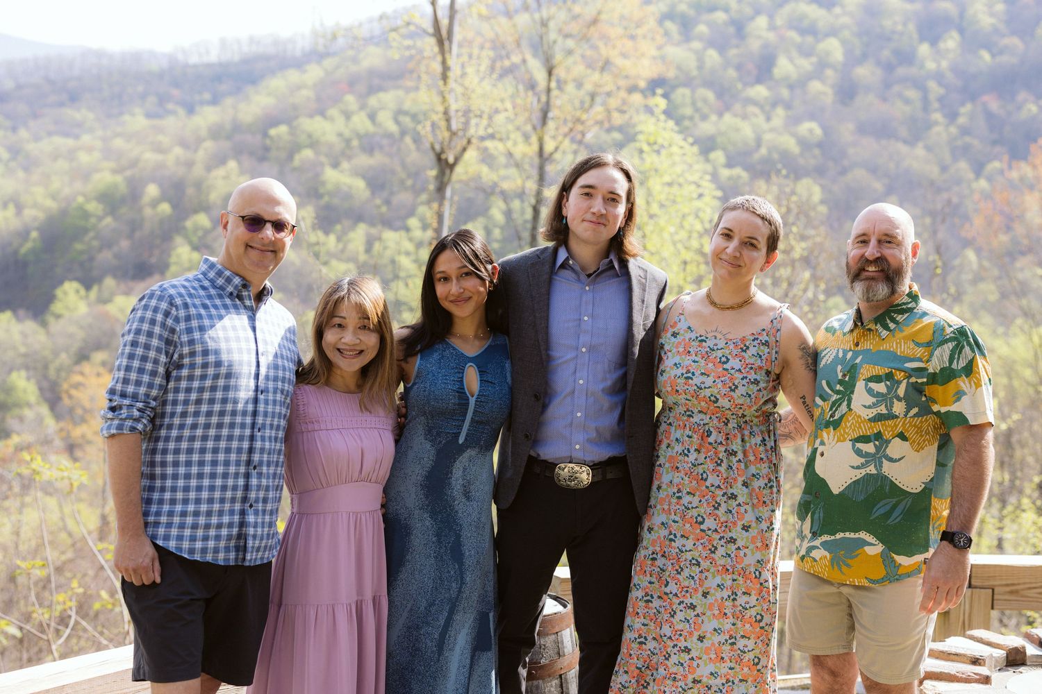 Group of six people smiling together on an outdoor deck with colorful autumn mountain foliage behind them.