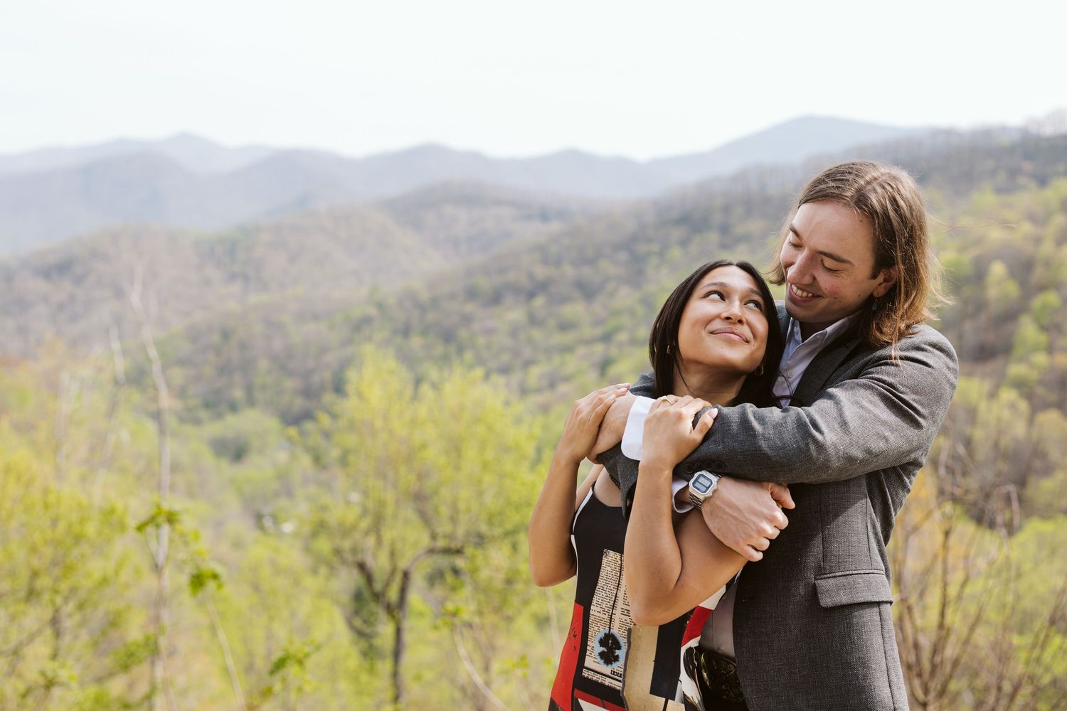 Happy couple embracing outdoors with woman smiling up at partner against a stunning green mountain landscape.