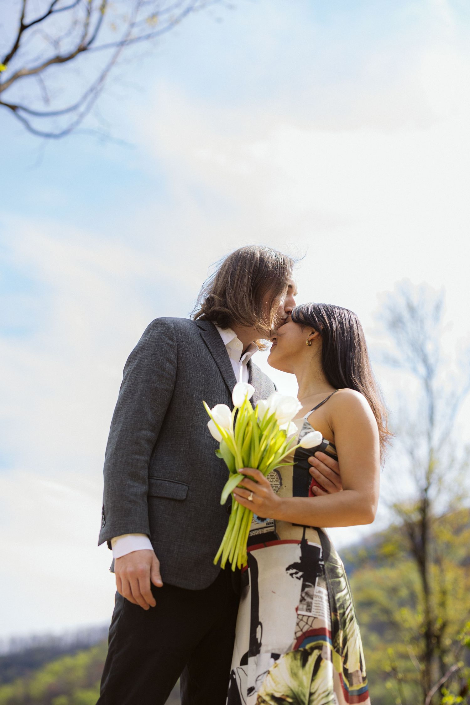 Couple sharing a romantic kiss while woman holds white tulip bouquet with blue sky and trees behind them.
