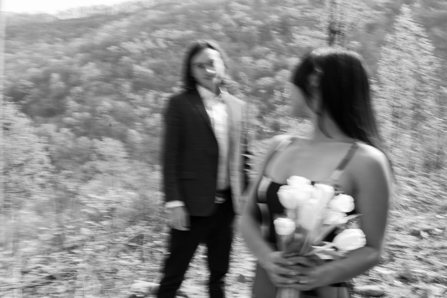 Black and white photo of a couple in nature, woman holding white flowers while man gazes at her.