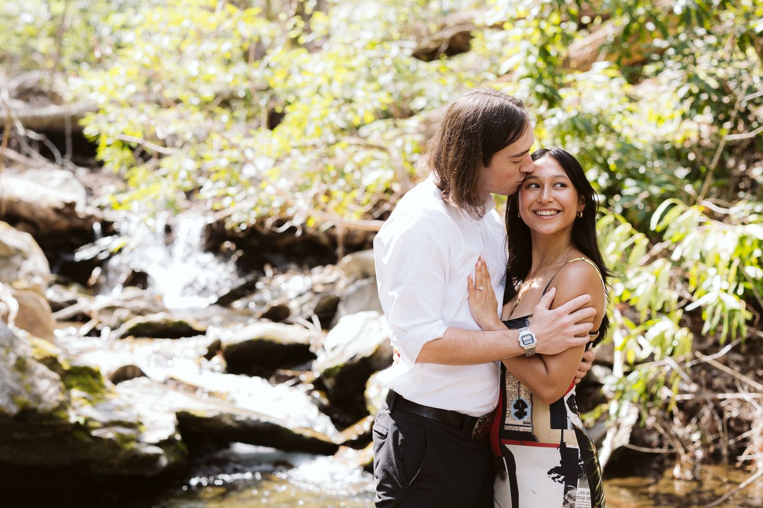 Couple embracing and kissing by a rocky waterfall surrounded by lush green trees.