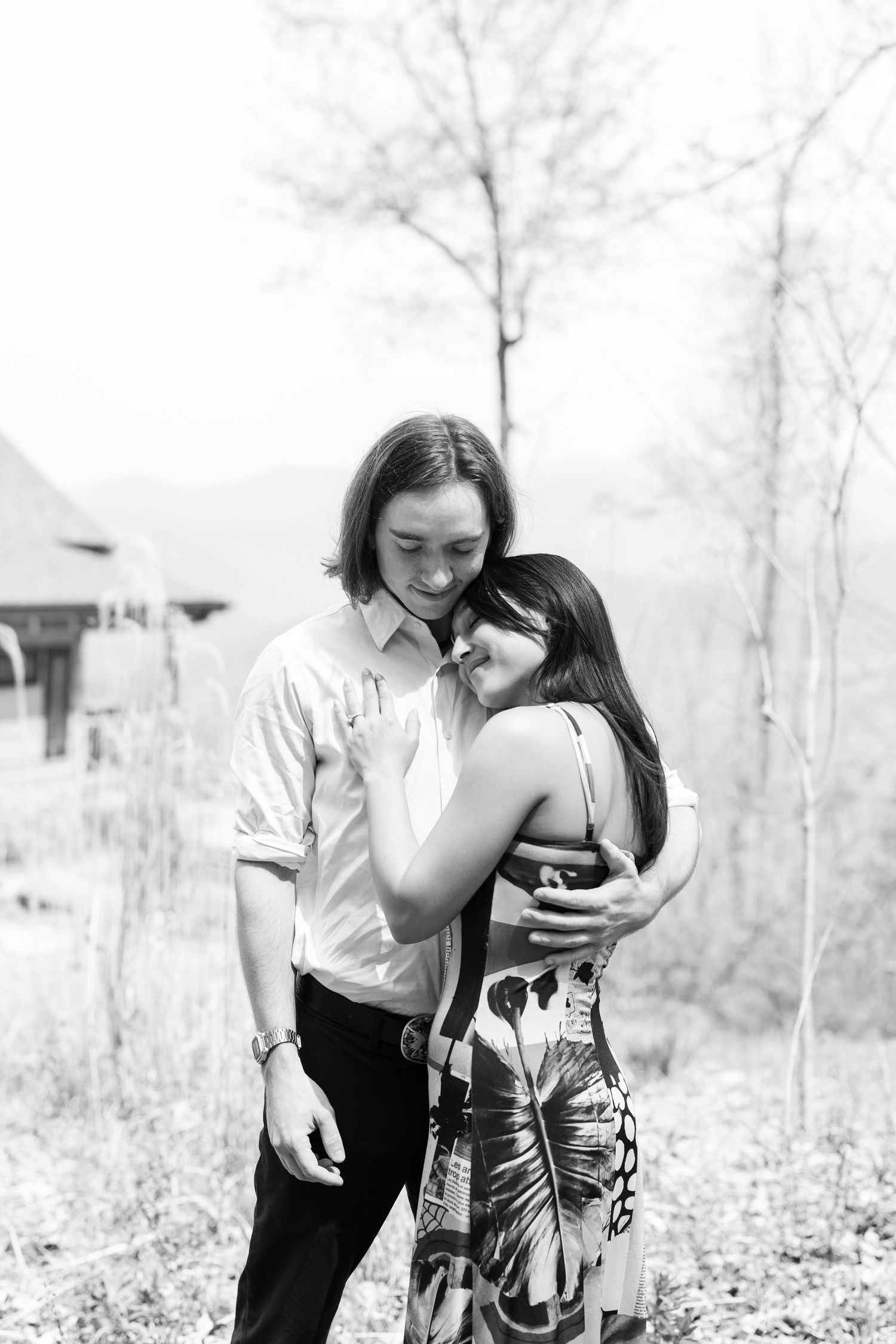 Black and white photo of couple embracing tenderly outdoors near rustic building.