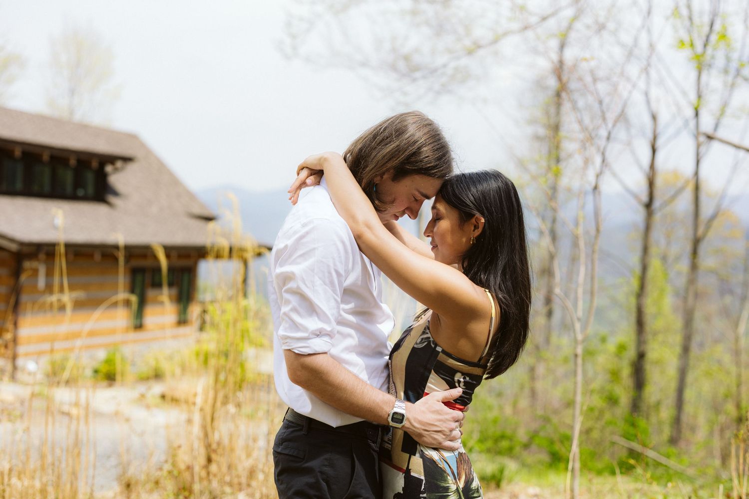 Couple sharing a romantic moment outdoors with rolling hills and rustic building behind them.
