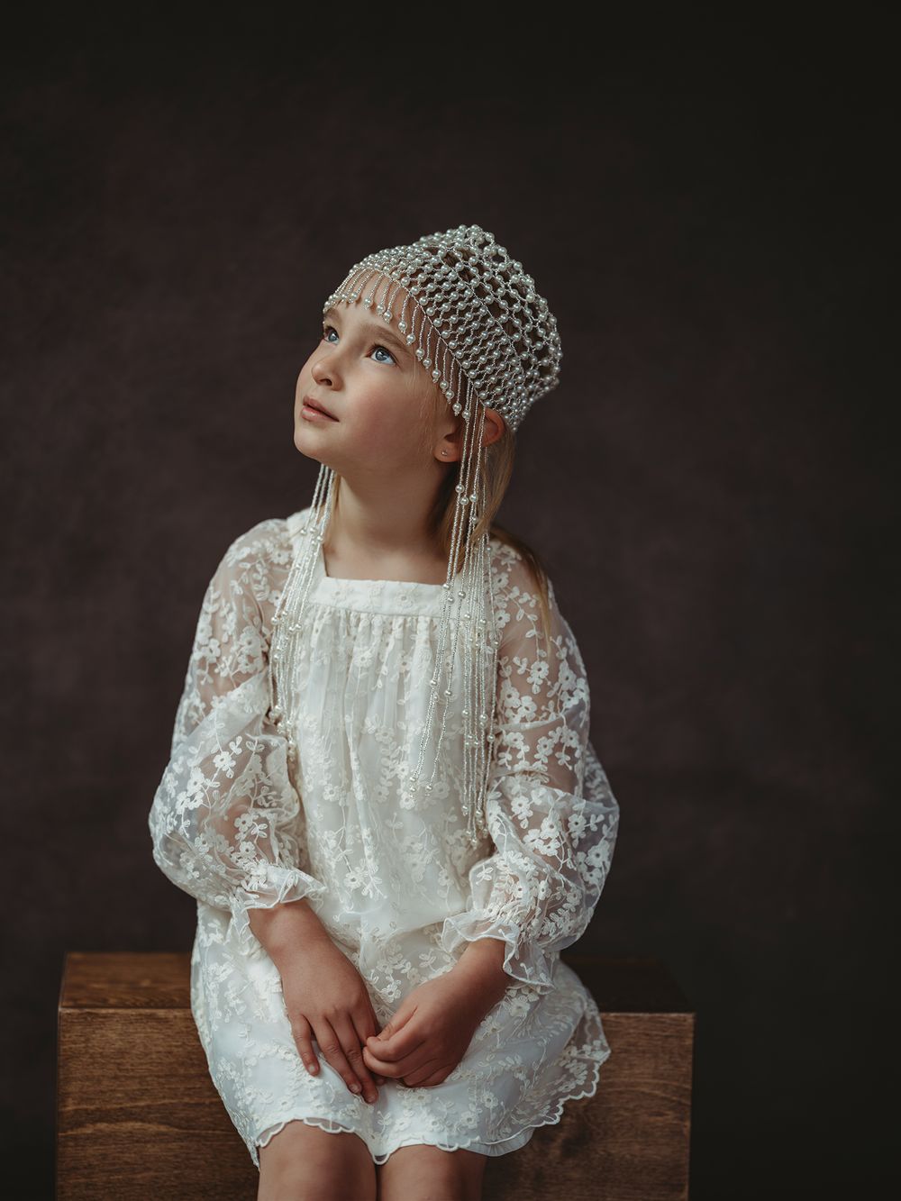 A baby in a white lace dress and knit cap sits on a wooden bench against a dark background.