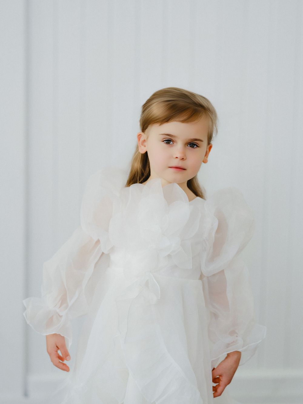 Young girl in elegant white organza dress with puffy sleeves and angel wings, posing gracefully against a white background.