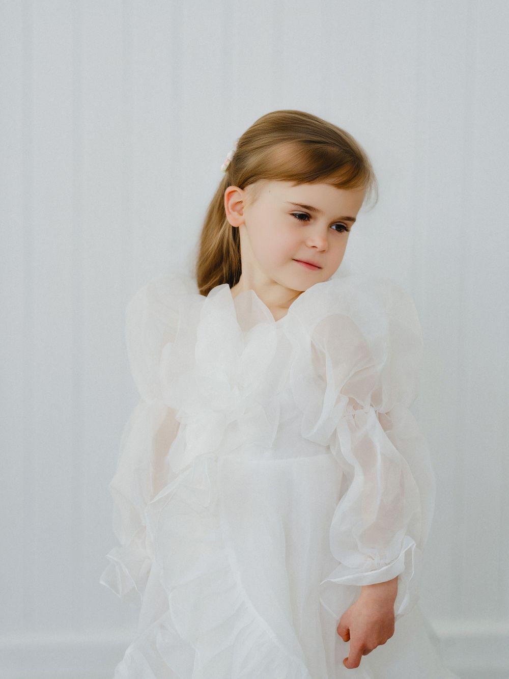 Young girl in white organza dress with puffy sleeves poses gracefully against a light background.