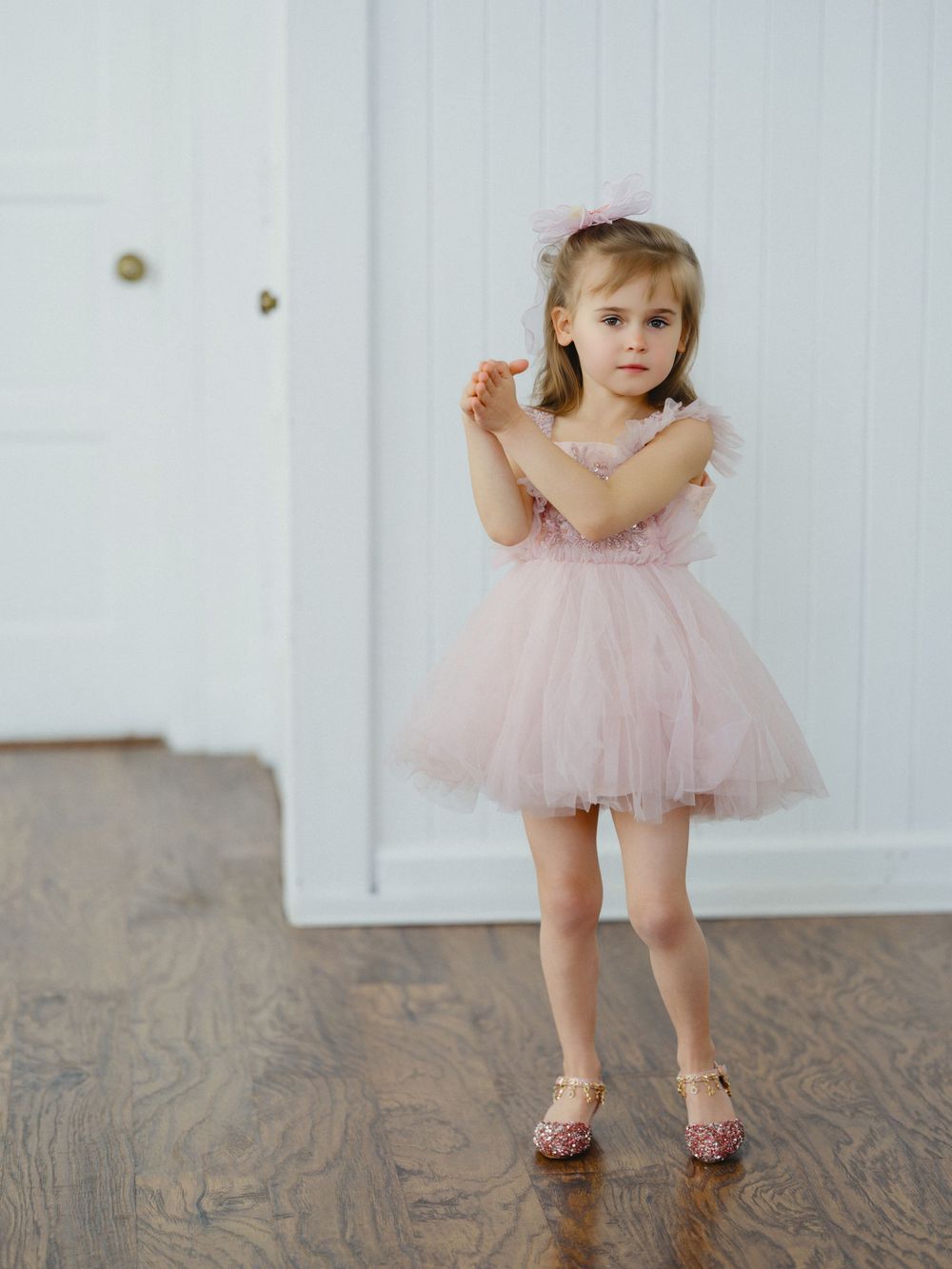 Toddler girl in pink tulle dress and glitter shoes stands on hardwood floor wearing floral headband.
