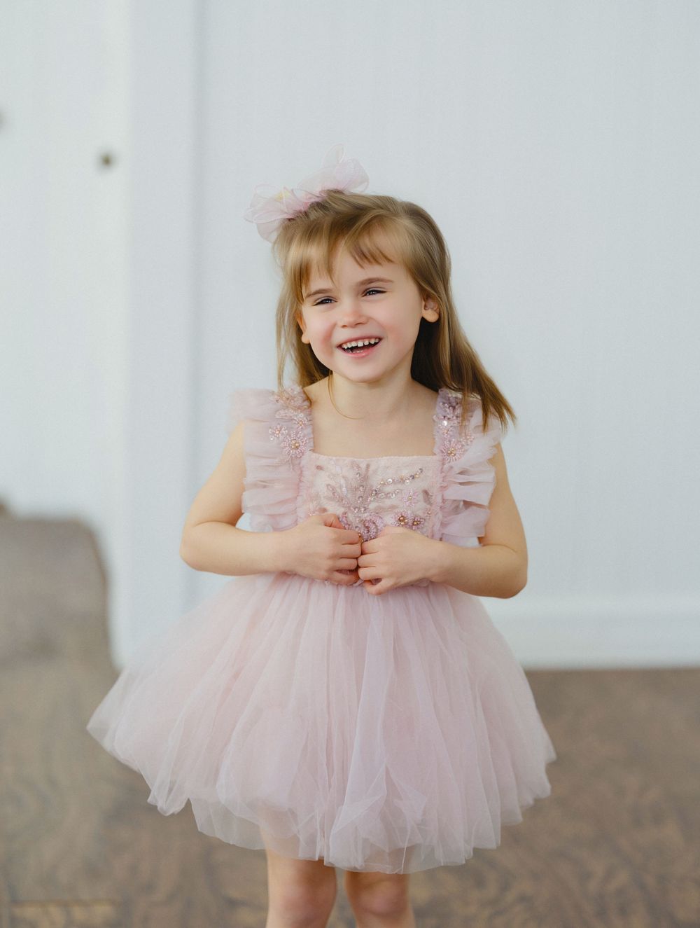 A smiling toddler girl wearing a pink tulle dress with ruffled straps and a matching bow hair clip.