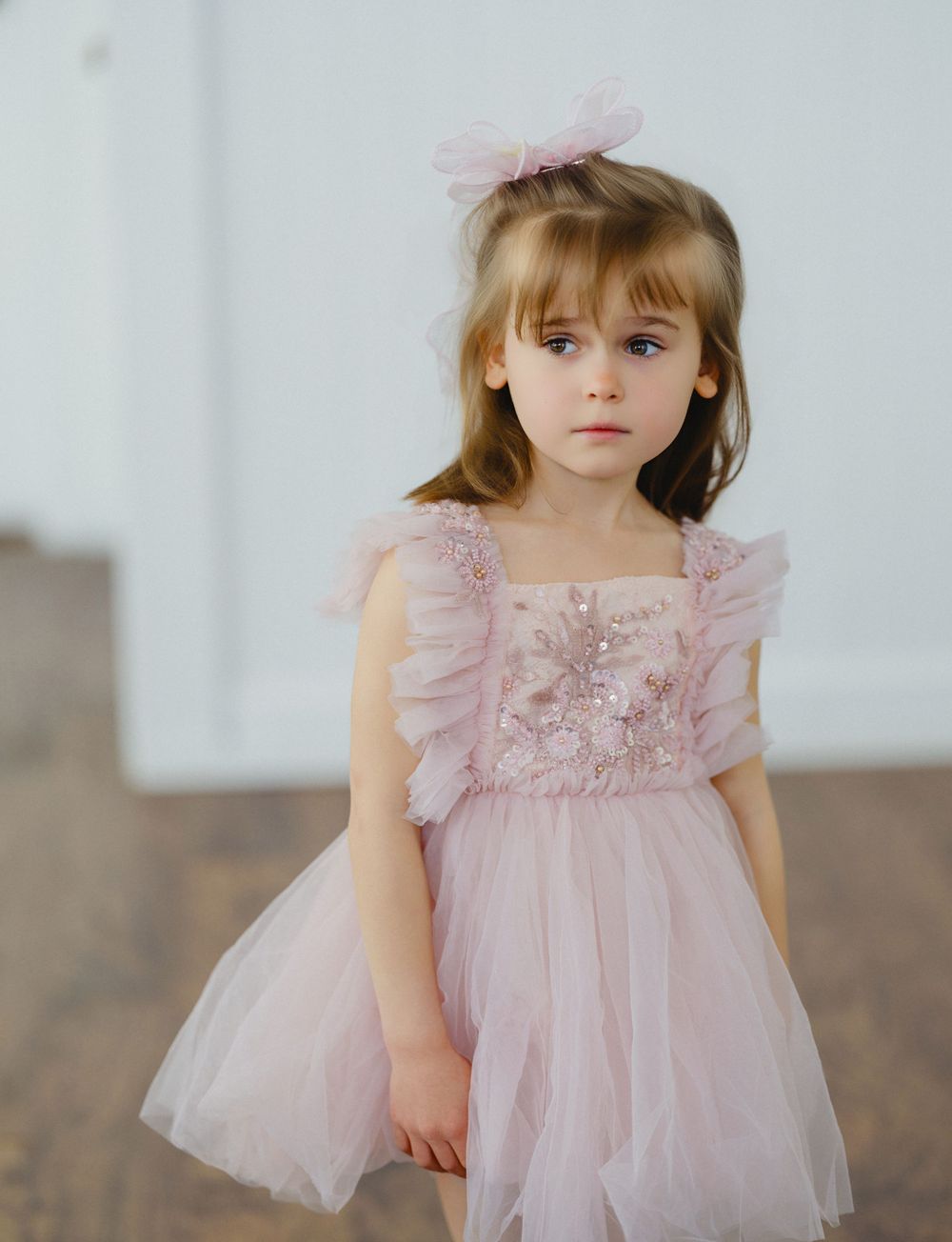 Young girl in pink tulle dress with floral embroidery and bow headband poses gracefully indoors.