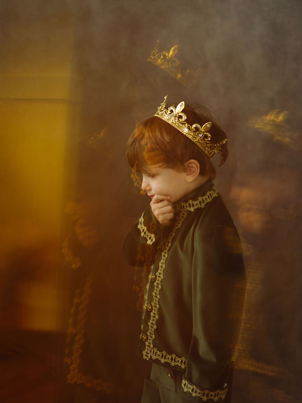 A red-haired child wearing a gold crown and dark royal costume poses thoughtfully in warm golden light.