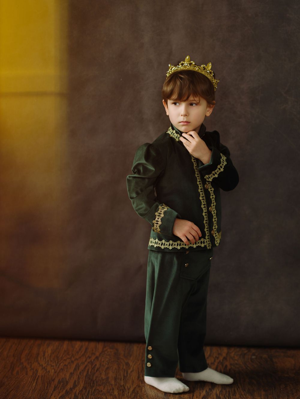 Young boy in green and gold prince costume with crown poses regally for portrait photo.