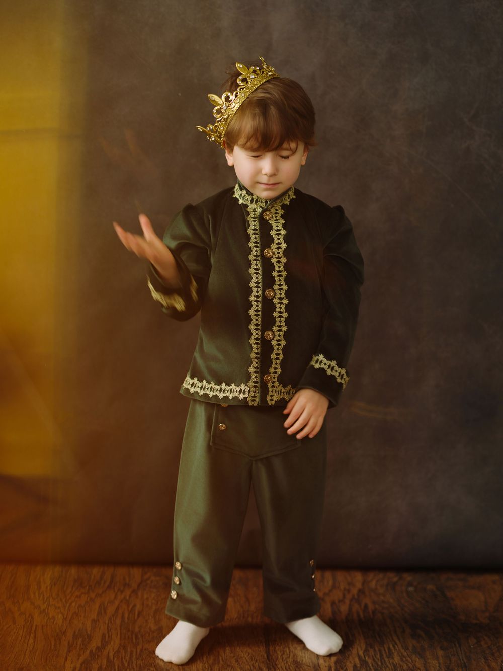 Young boy dressed in dark green and gold royal costume with crown, posing regally in a studio setting.