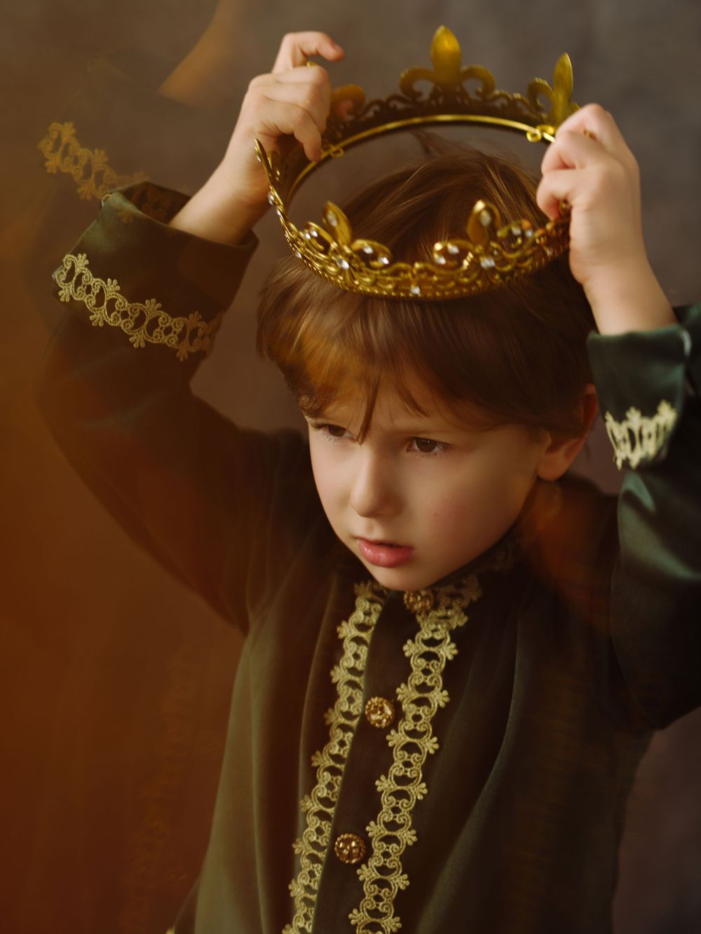 Young boy in royal medieval costume placing a gold crown on his head against a dark background.