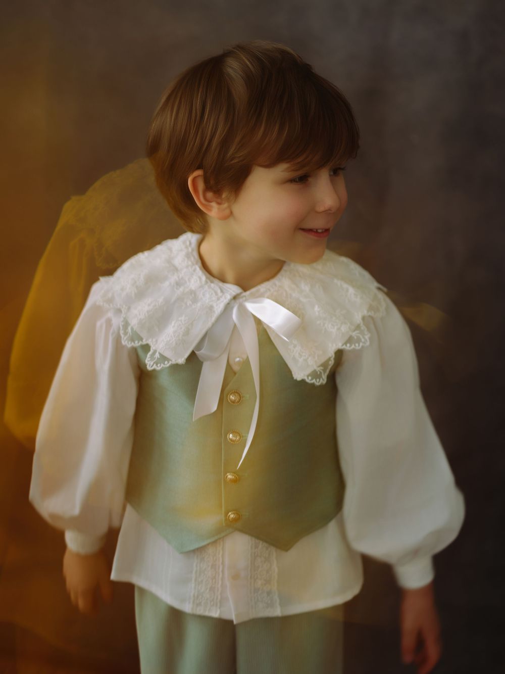 Young boy in vintage-style sage green vest, white lace collar blouse, and trousers, smiling softly.
