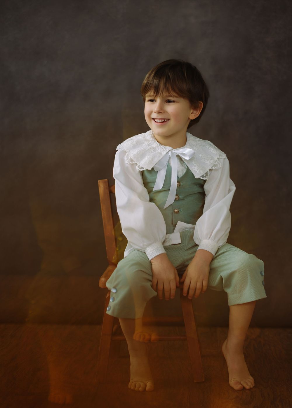Smiling young boy in vintage green vest and white ruffled blouse seated on wooden chair for portrait.