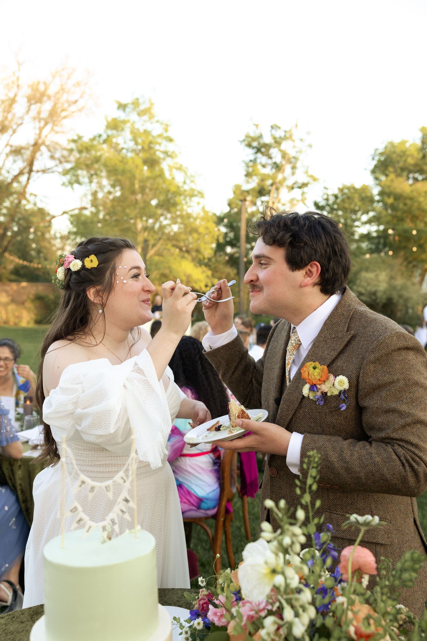 Newlyweds playfully feeding each other cake at outdoor wedding reception.