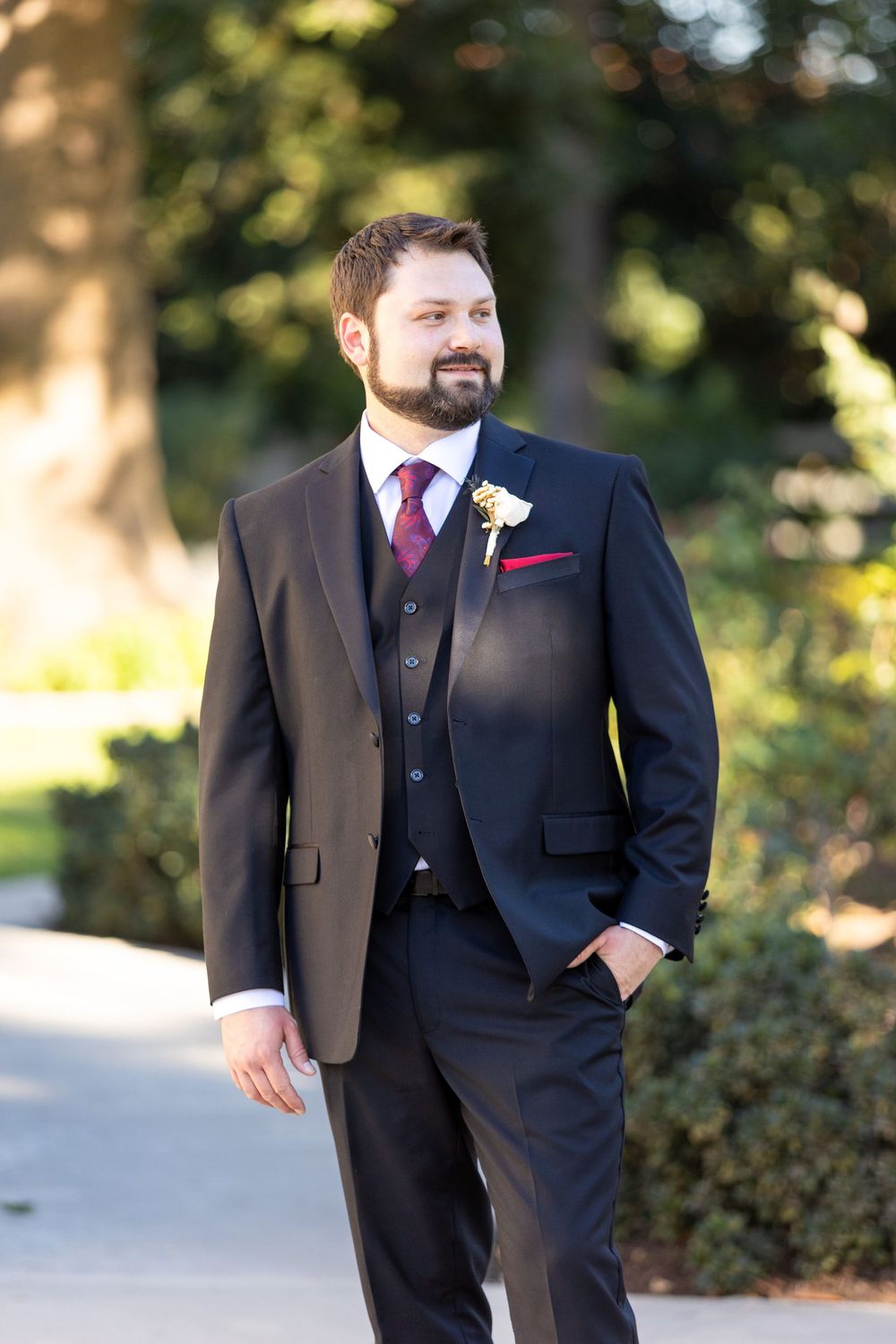 Groomsman in a dark suit with a pink tie stands outdoors in natural lighting during a wedding.