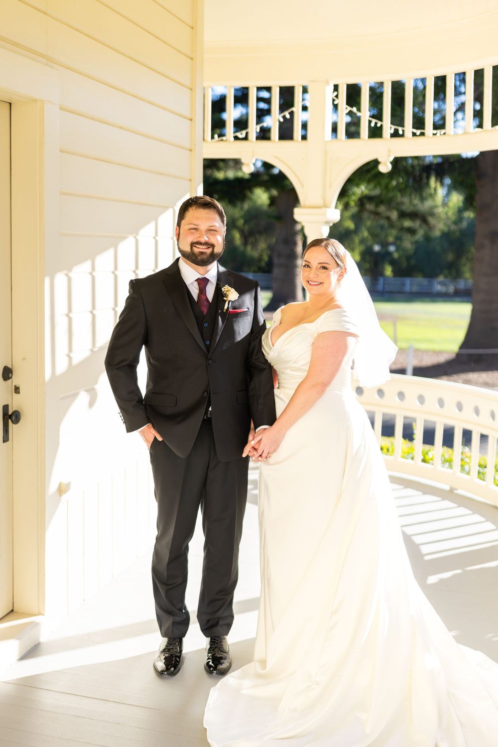 A couple in formal wedding attire poses together in a white gazebo with sunlit columns.