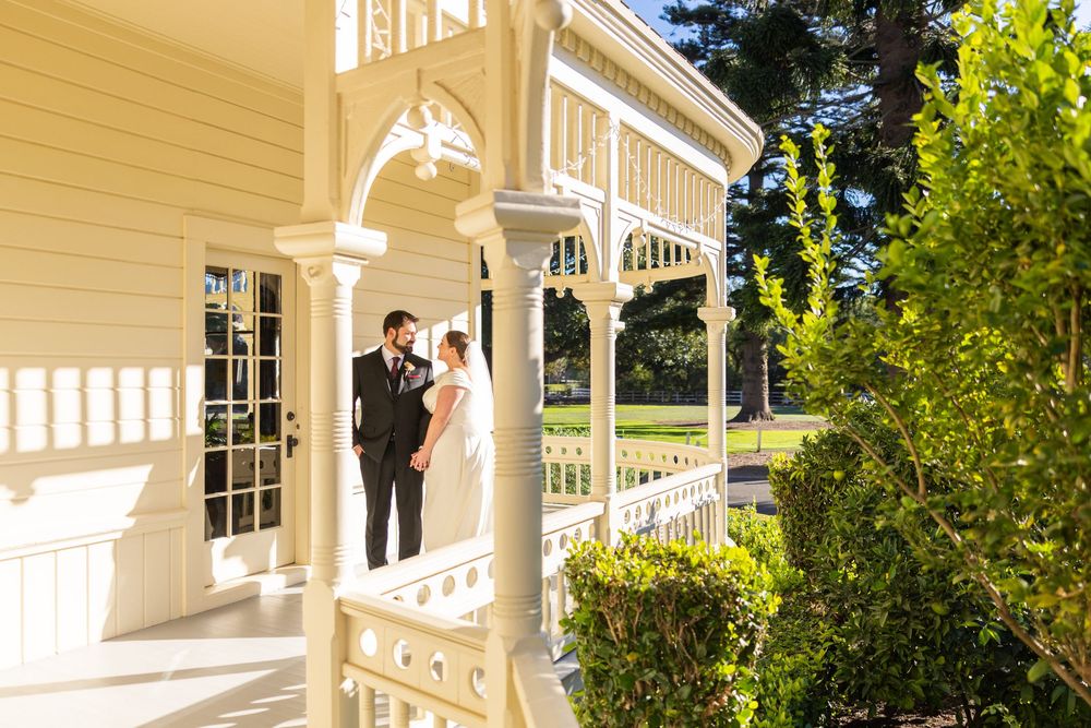 White Victorian-style porch with ornate trim and columns surrounded by lush green foliage on a sunny day.