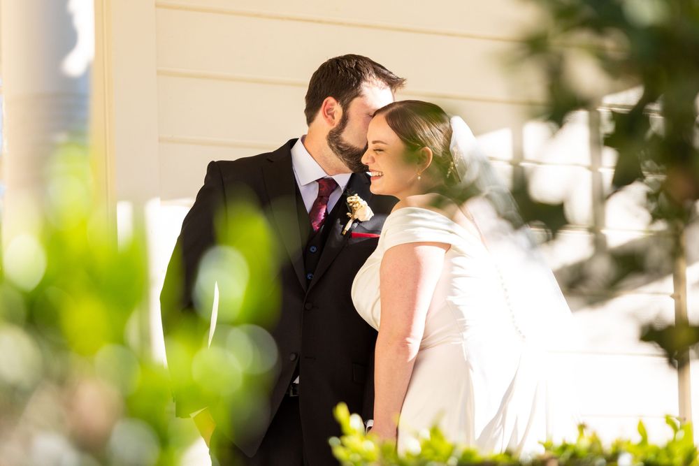 A romantic wedding moment captured between a couple in formal attire standing close together among green foliage.
