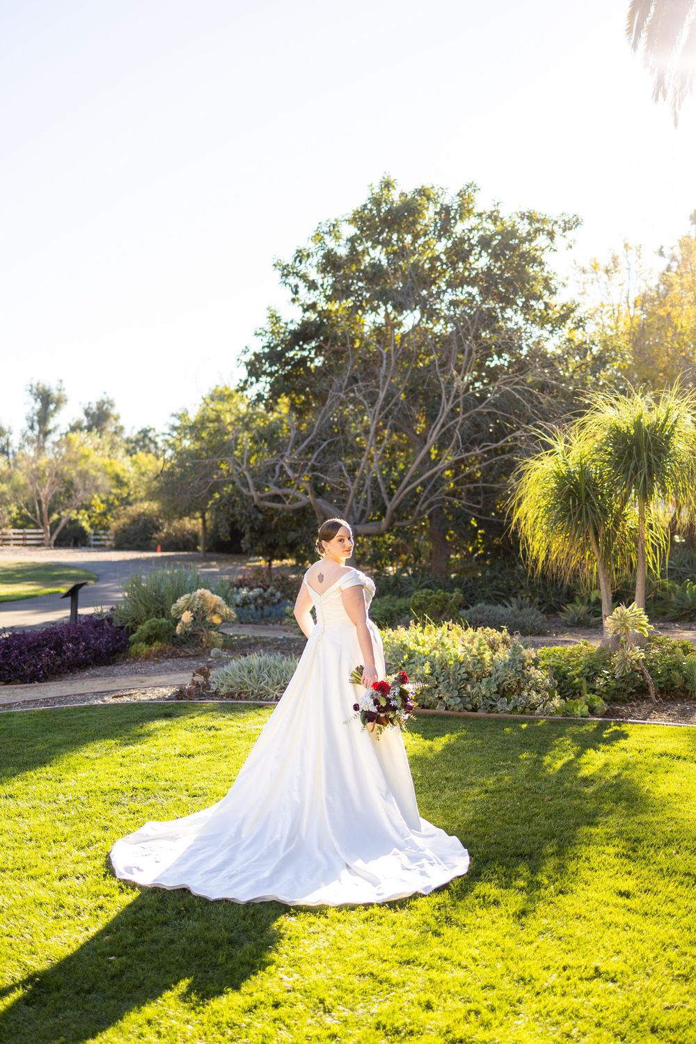 Beautiful wedding dress flows across sunlit green lawn in garden setting.