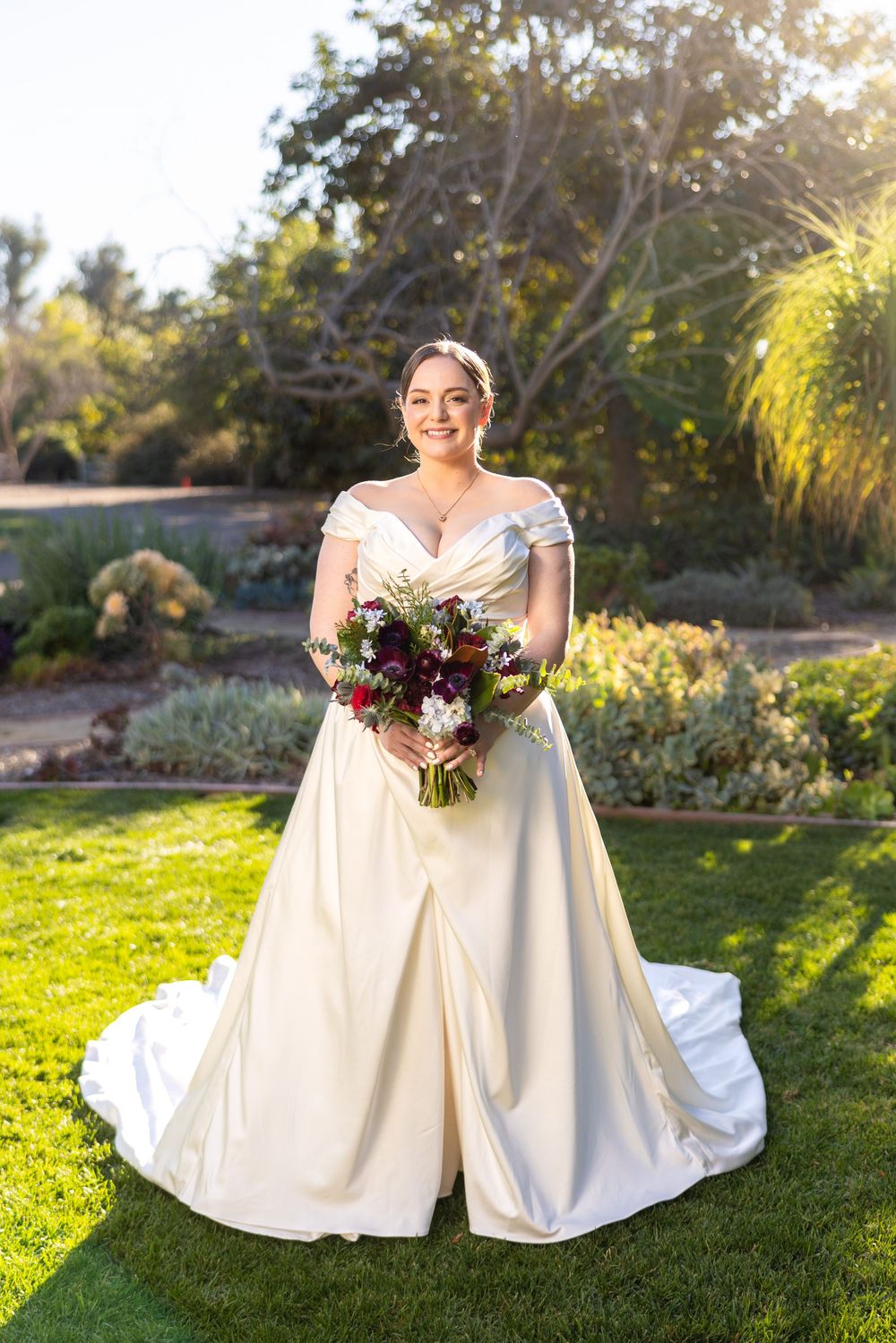 Bride in white wedding dress sits gracefully on grass holding vibrant burgundy and white bouquet at sunset.