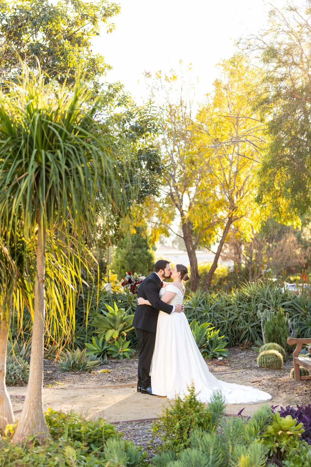 Wedding couple shares intimate moments in a lush garden setting with palm trees and golden sunlight.