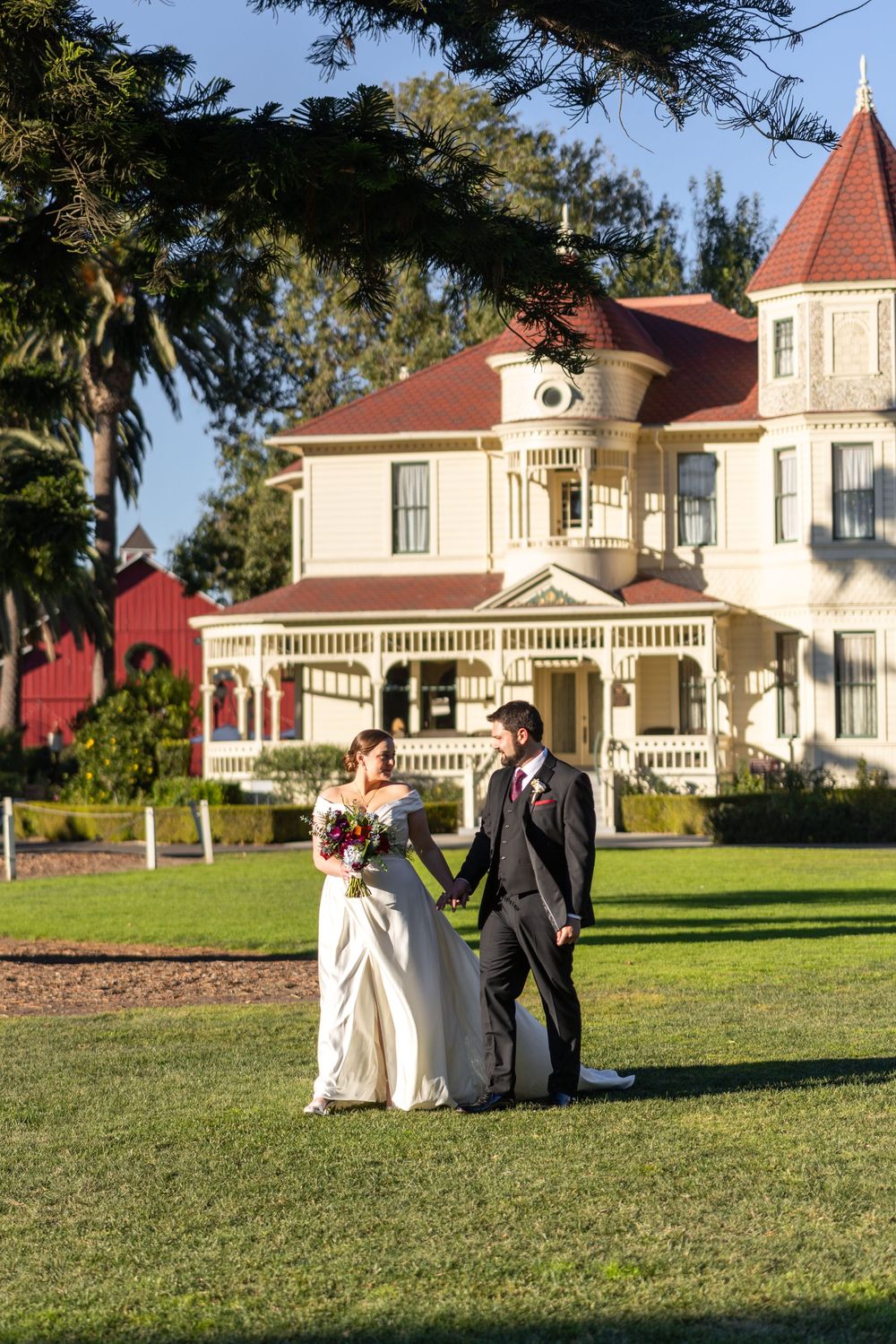 A couple poses for wedding photos on a grassy lawn in front of Victorian-style buildings with red roofs.