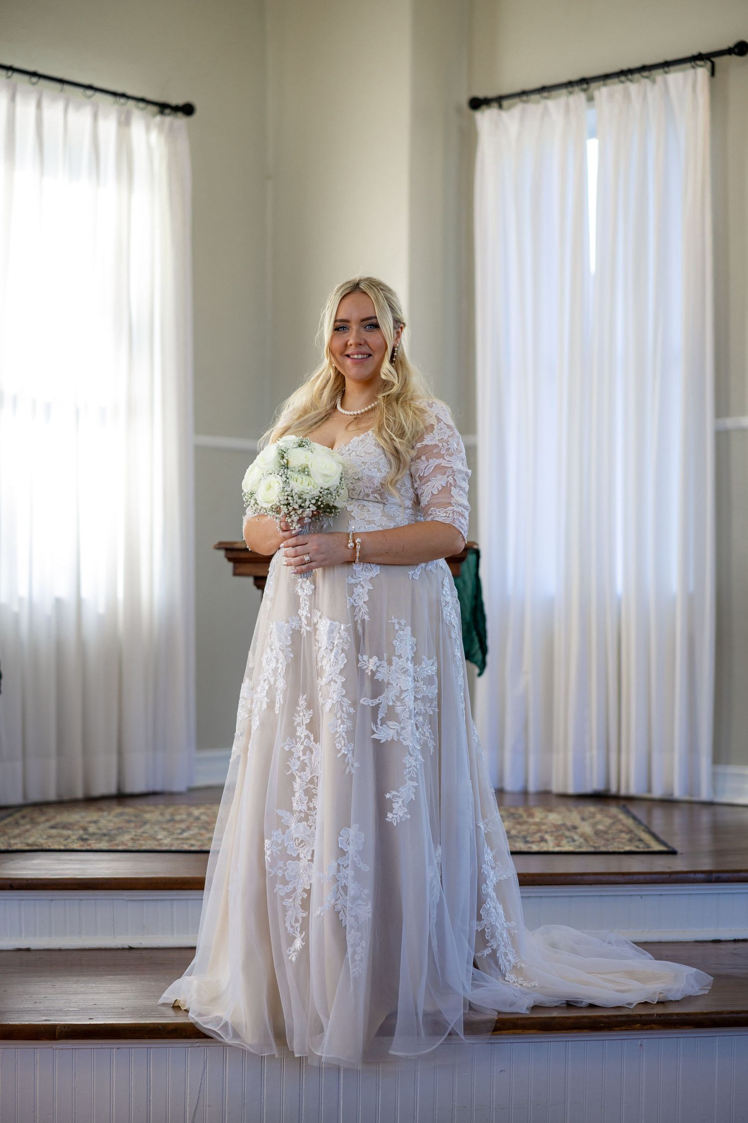 Smiling bride in lace wedding gown holding white bouquet stands on stairs before large windows.