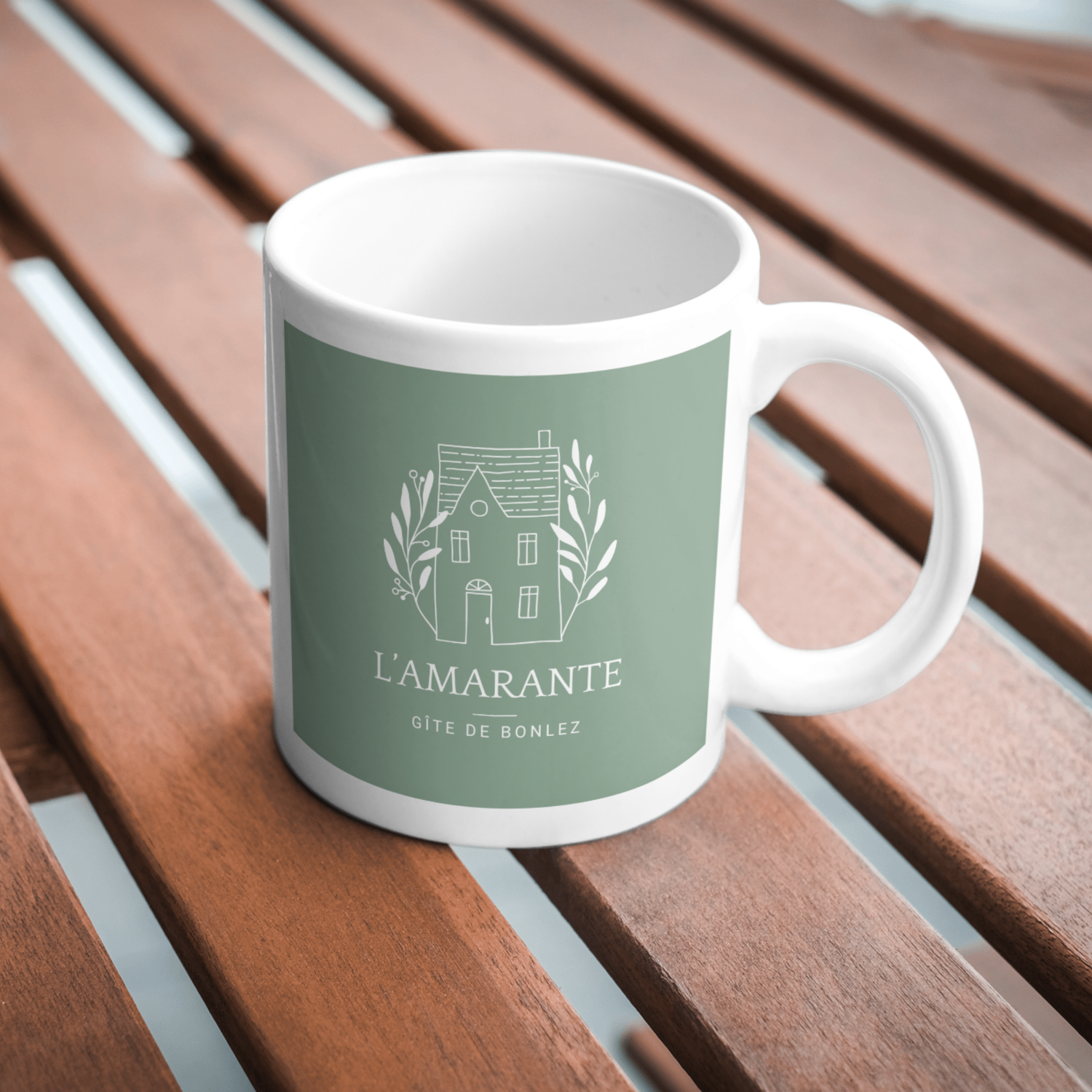 White ceramic coffee cup with green L'amarante branding shown from multiple angles on wooden slat surface.