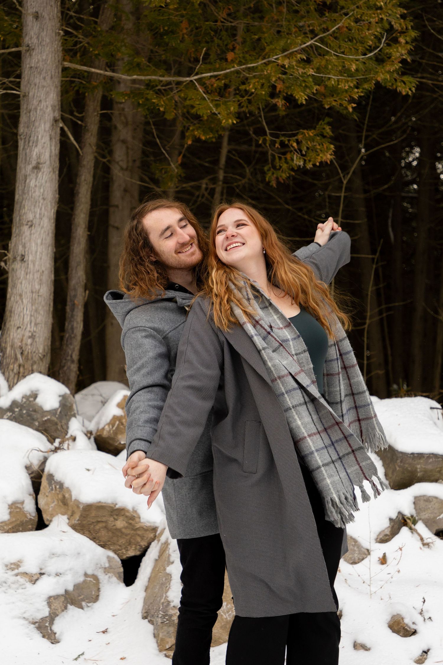 A Couple in the winter outside with snowy rocks behind them holding hands with their arms stretched out wide