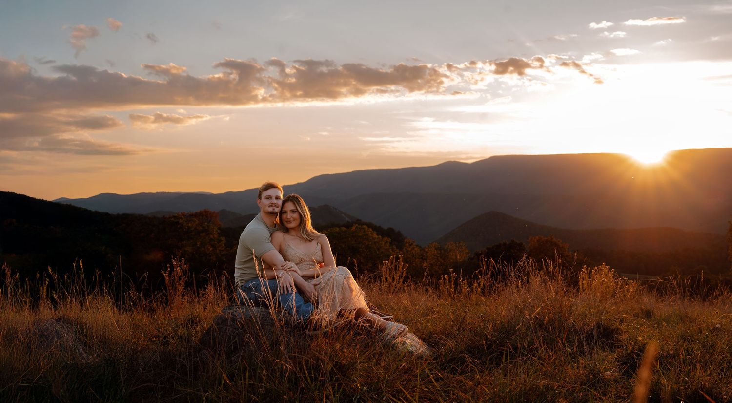 A couple sits together on a grassy hilltop at sunset with mountains silhouetted in the background.