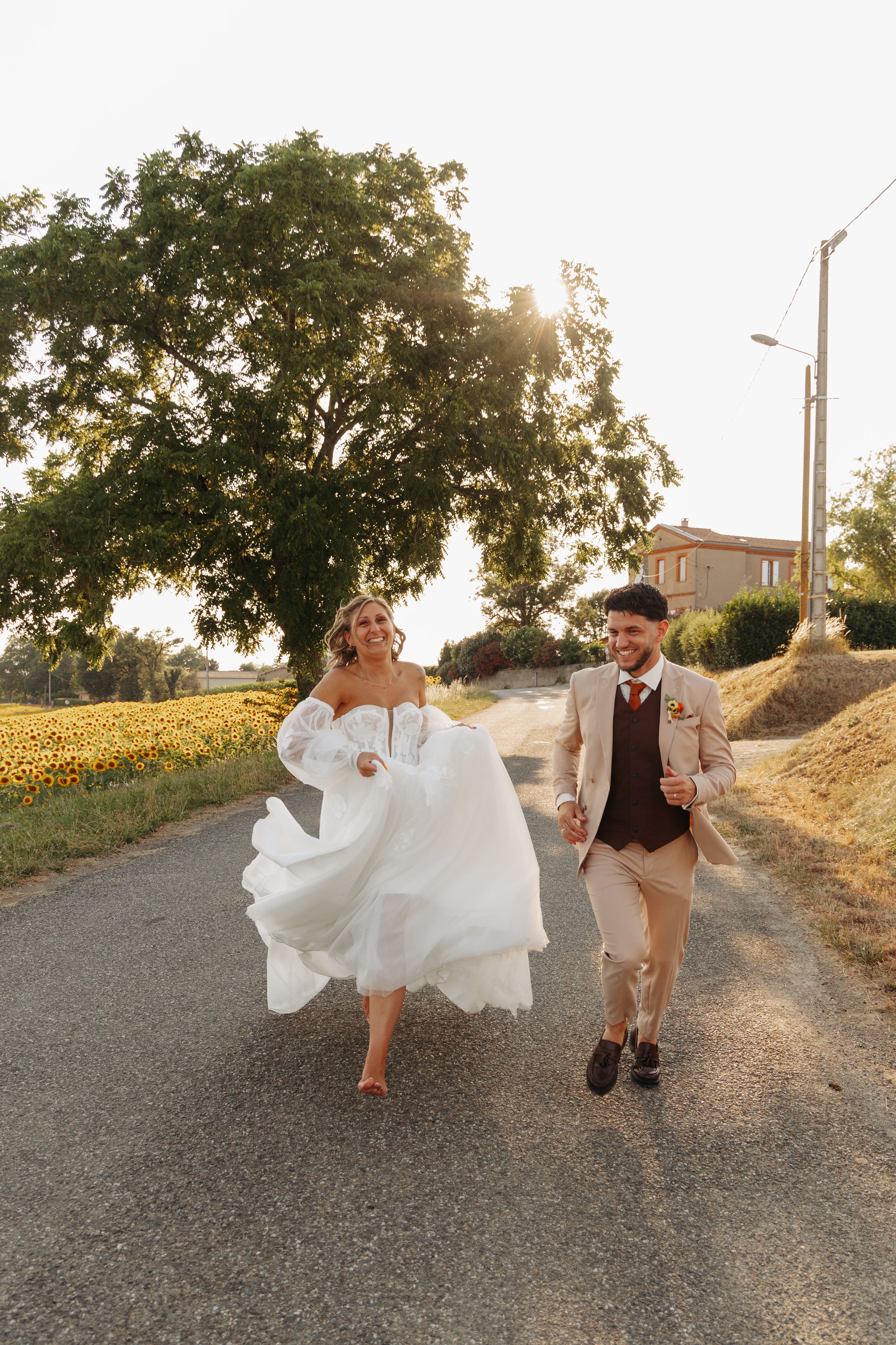 Bride and groom running hand in hand down a sunlit country road near sunflower fields.