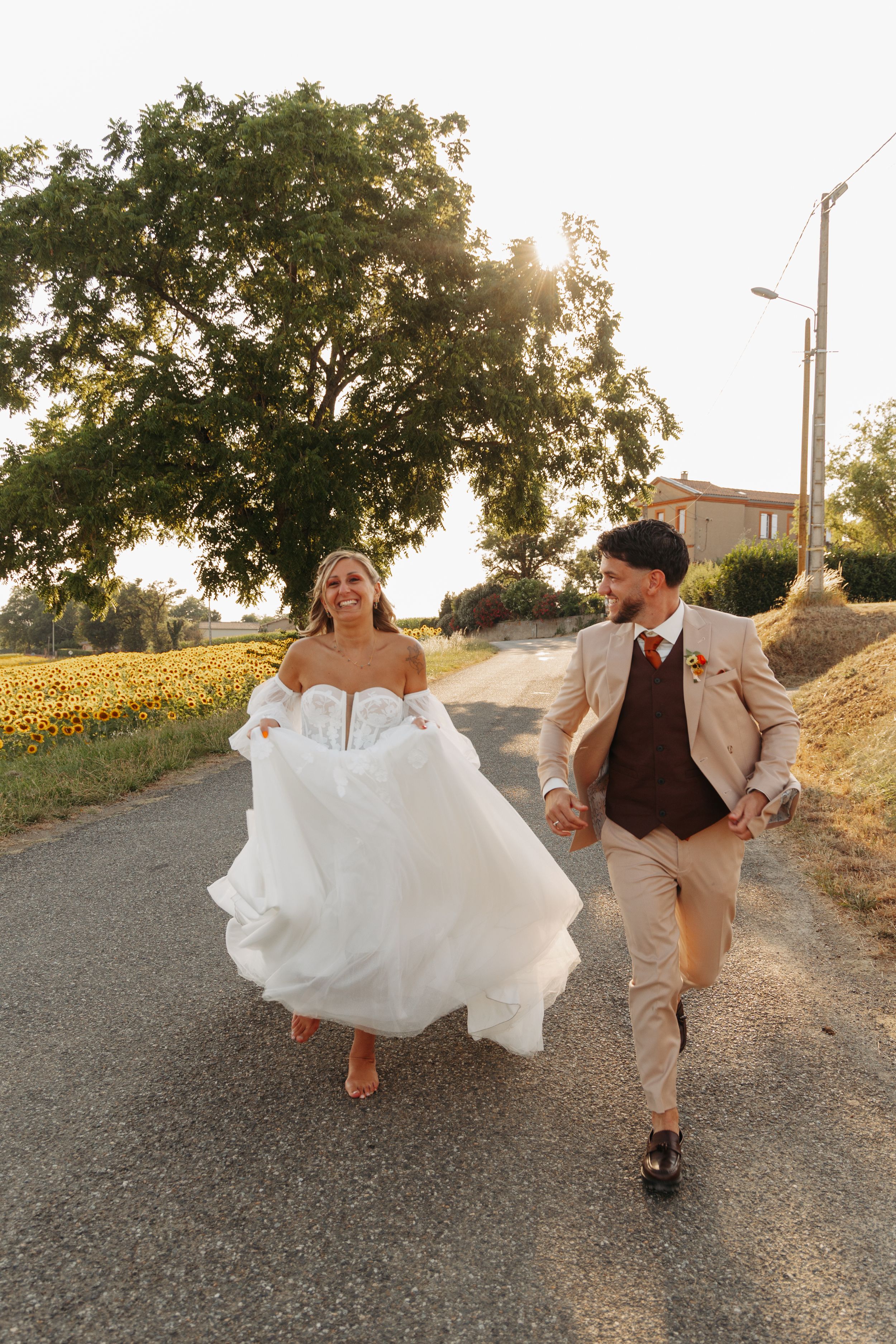 Bride and groom holding hands running down a sunny road near sunflower fields.