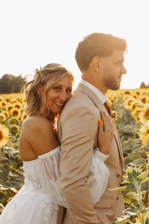 Smiling bride in white off-shoulder gown leaning against groom in beige suit in a sunflower field.