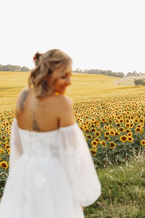 Bride in white off-shoulder wedding dress with tattoo standing in a blooming sunflower field.