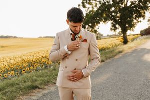 Groom in beige double-breasted suit adjusting boutonniere on a countryside road near sunflower fields.