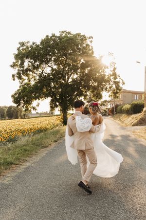 Groom carrying bride in white dress down a sunlit country road beside a sunflower field.