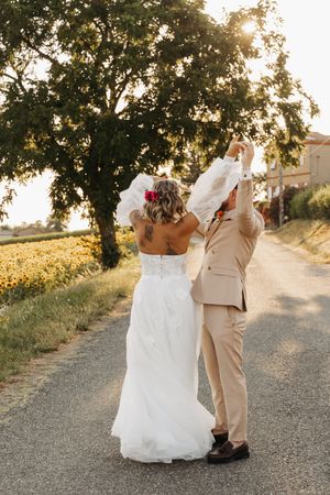 Bride and groom dancing joyfully on a rural road with sunflower fields and golden sunset backdrop.