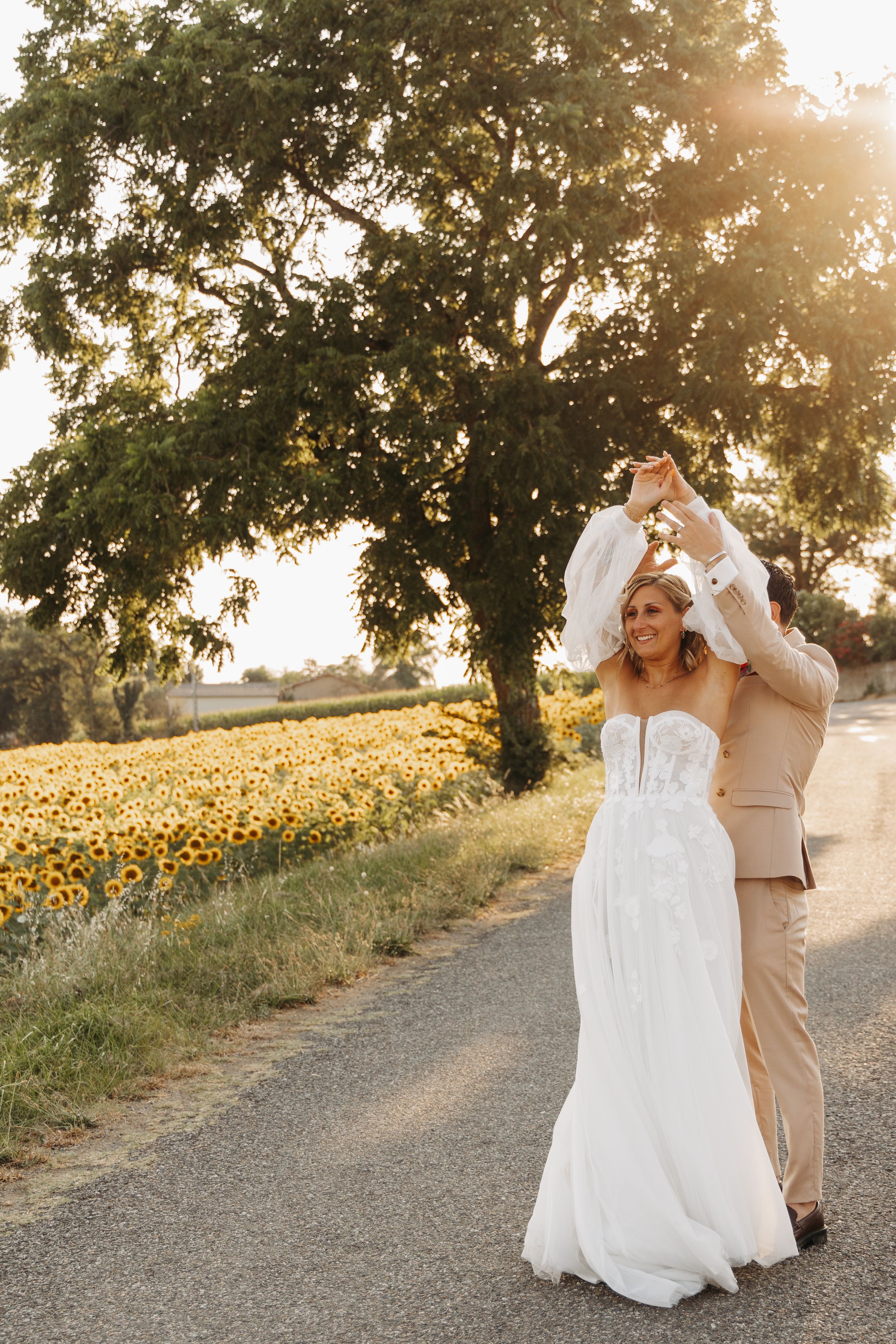 Groom twirling bride in white wedding dress on a countryside road beside a sunflower field at sunset.