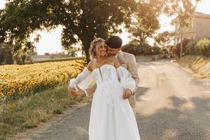Groom in beige suit kissing bride in white lace gown on a sunny road by sunflowers.
