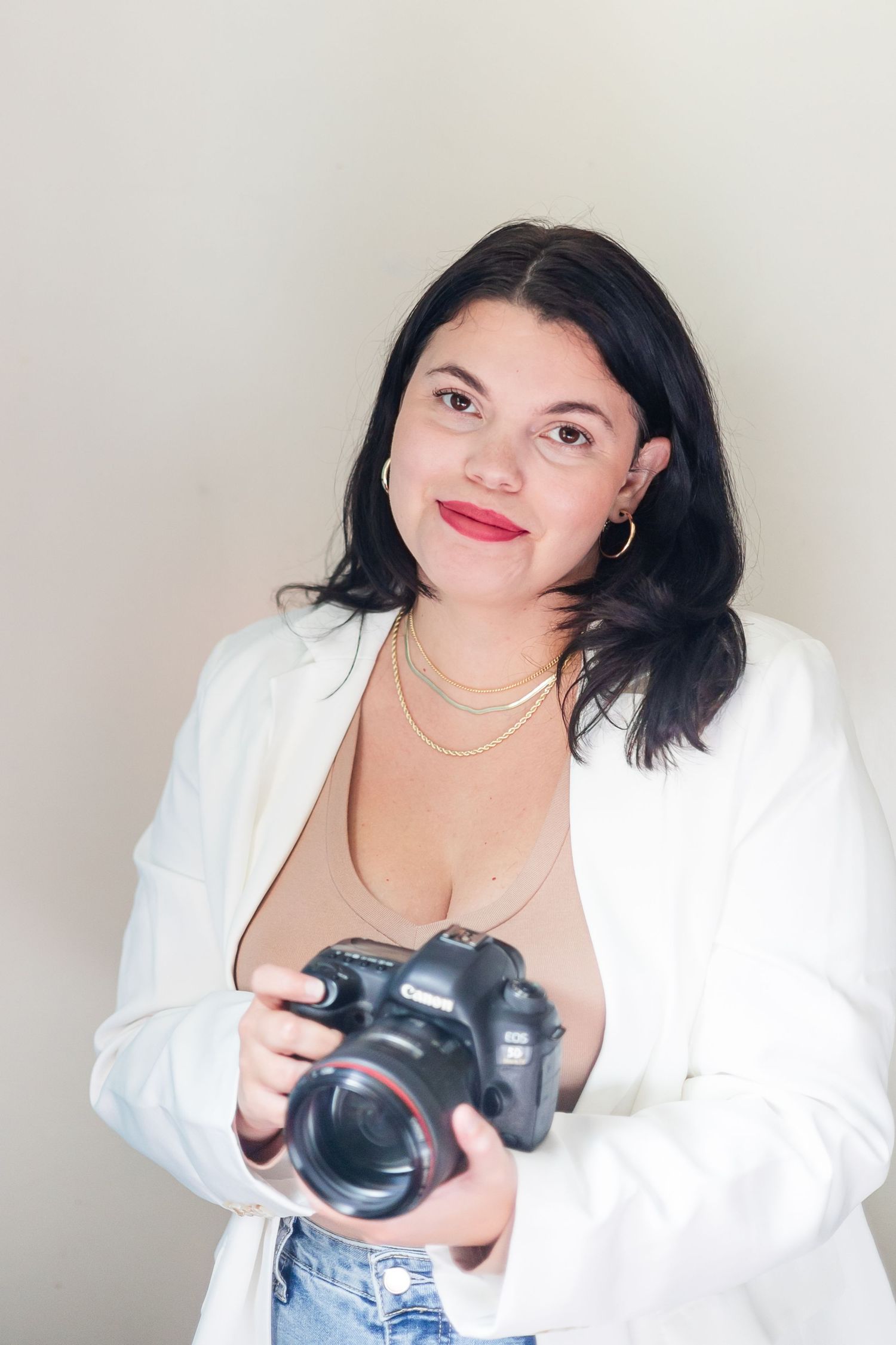 Professional photographer in West Virginia white blazer and jeans holding DSLR camera against neutral background.