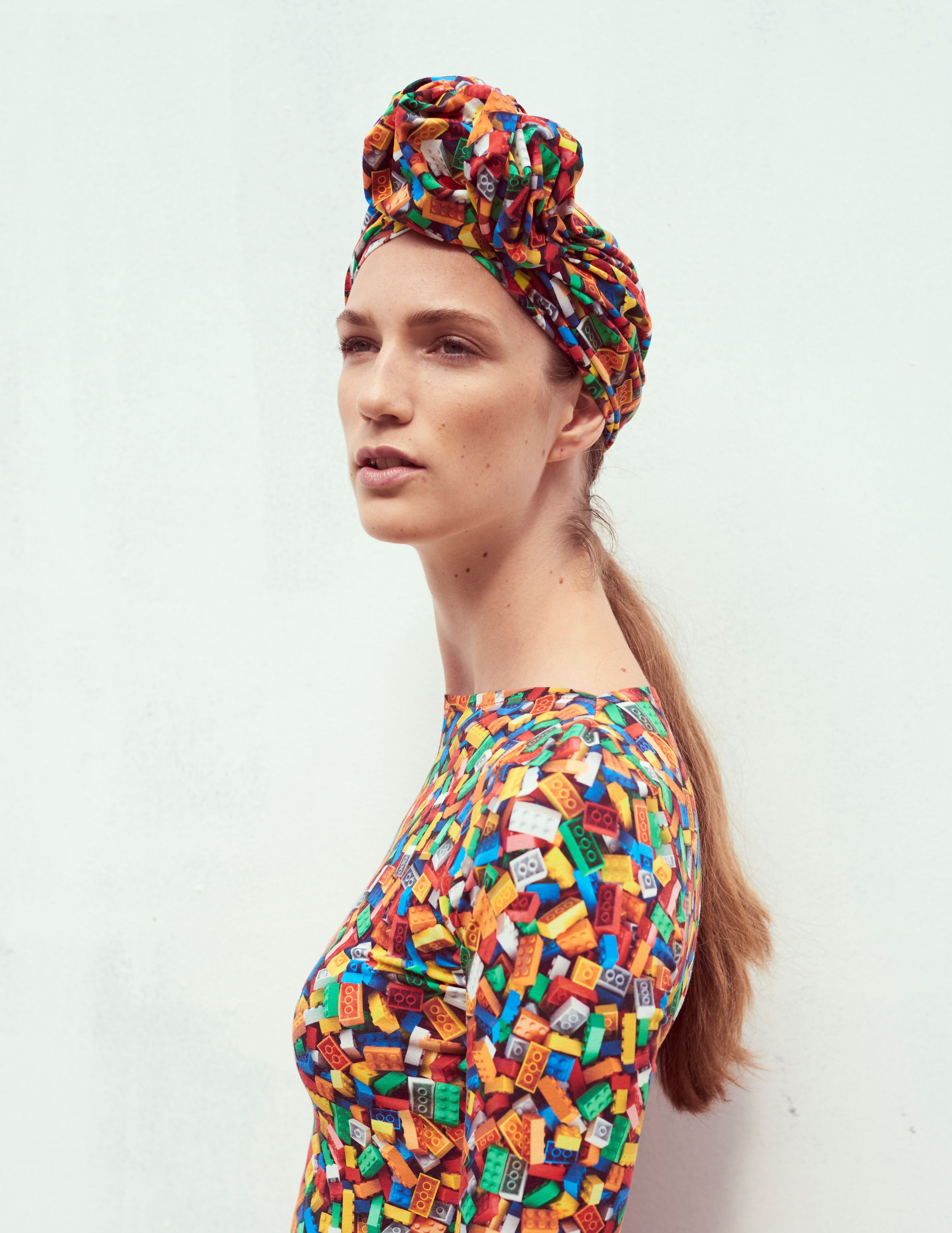 Fashion model wearing a colorful printed dress and matching turban headwrap against a white wall.