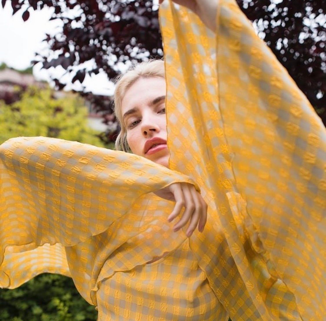 Blonde woman wrapped in yellow gingham fabric outdoors with green trees in background.